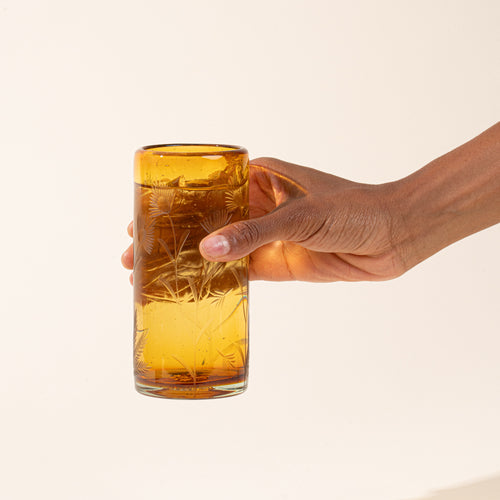 A hand holding a tall, cylindrical amber glass with etched floral patterns, set against a plain off-white background.