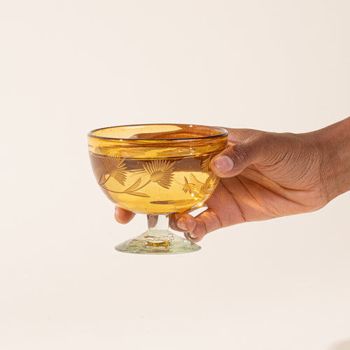 A hand holds an amber etched coupe glasses with a pedestal base and delicate botanical pattern, filled with liquid.