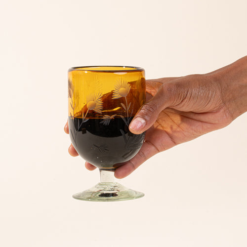 A hand holds an amber short wine glasses made of recycled glass with etched thistle designs on it, filled with red wine.