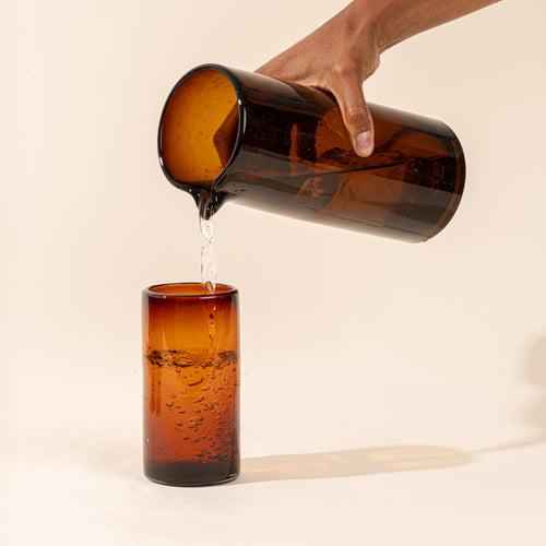 A hand holds a dark amber tall transparent glass pitcher with small spout at the top, pouring water into a matching tumbler.