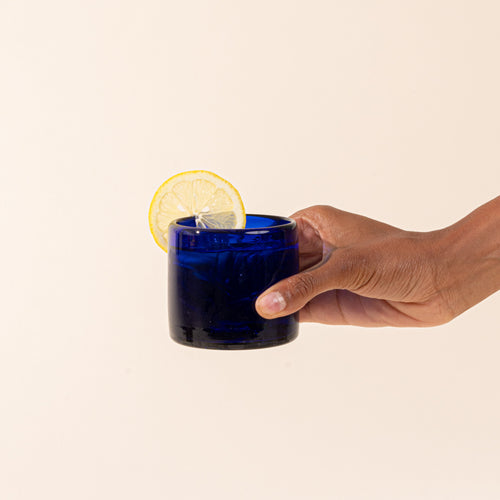 A cobalt blue short transparent drinking glass with a drink and lemon slice.