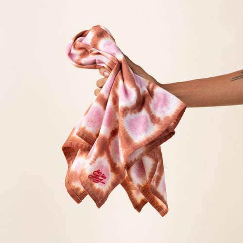 A hand holding out a tie-dye tea towel in a pink colorway with a red embroidered corner