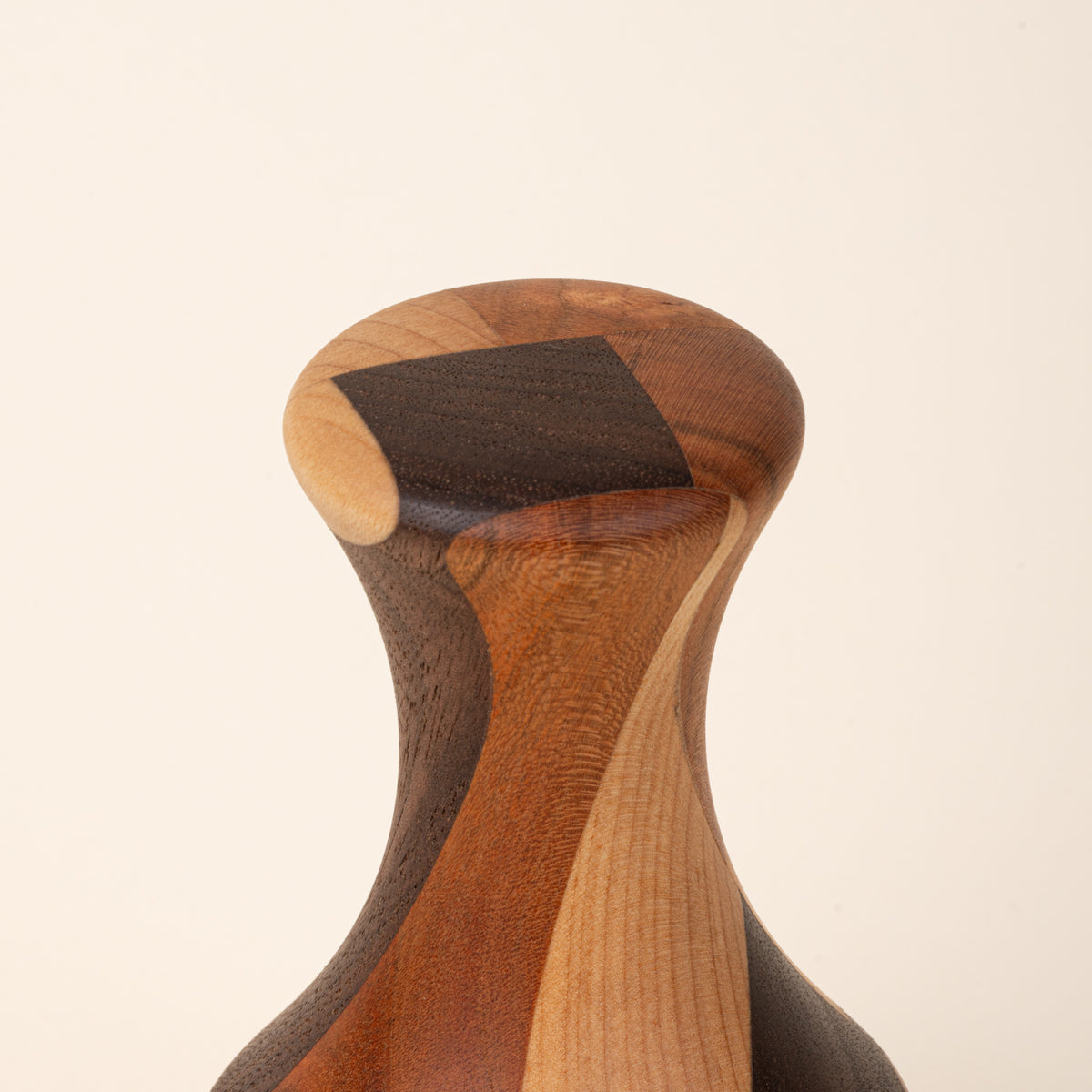 Closeup of a top of a Sculptural wooden biscuit cutter with a tapered neck, flared top, and geometric patchwork of light and dark wood tones.