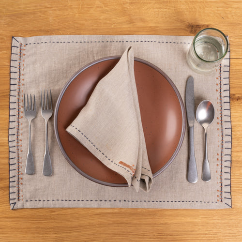 A natural linen placemat and coordinating napkin with dark blue running-stitch borders and rust-orange embroidered details, styled with a warm terracotta ceramic plate, silver flatware, and a clear glass on a wood tabletop.