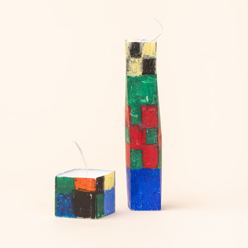 Two hand-painted geometric candles in coordinating designs: a short square pillar and a tall tapered rectangular candle. Both feature expressive blocks of cobalt blue, red, green, yellow, and black with white wicks and a textured, handmade finish.