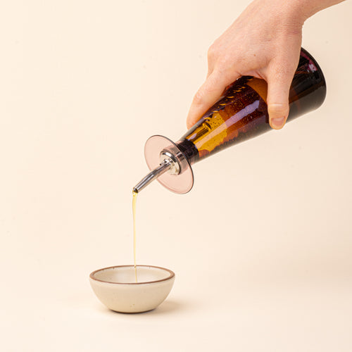A hand holds a plum glass oil or vinegar dispenser with a tapered bottle shape, wide flared collar, and metal pour spout with a black stopper, pouring oil into a small ceramic bowl.