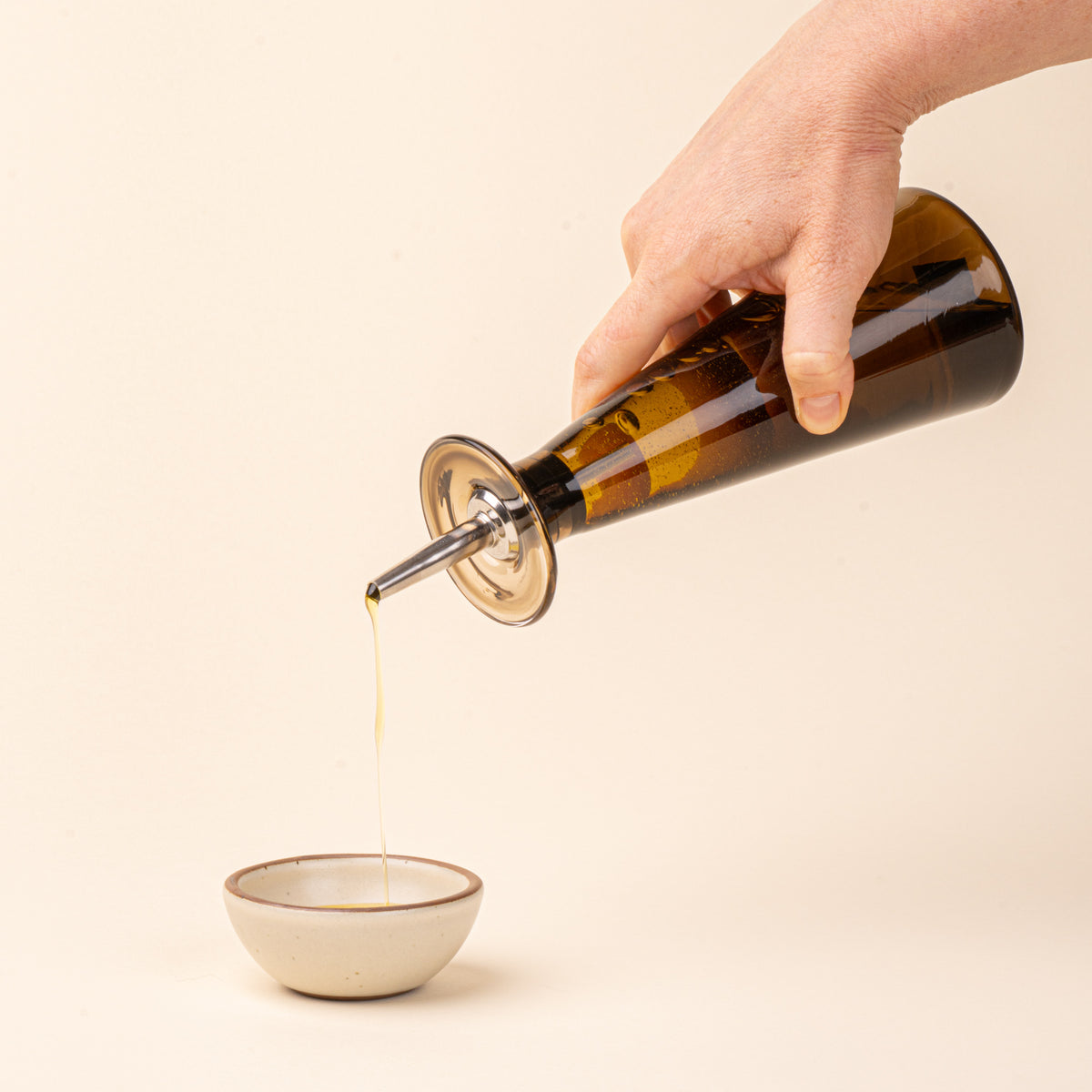 A hand holds an amber-brown glass oil or vinegar dispenser with a tapered bottle shape, wide flared collar, and metal pour spout with a black stopper, pouring oil into a small ceramic bowl.