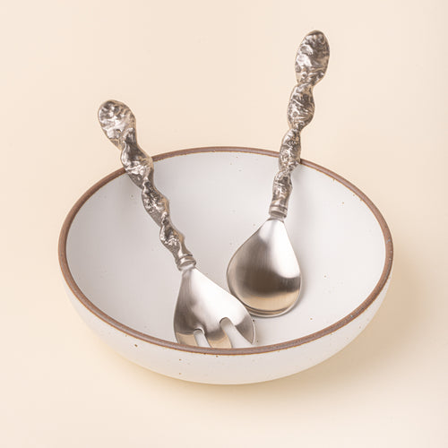 Stainless steel salad serving fork and spoon with textured twisted handles resting in a white ceramic bowl.