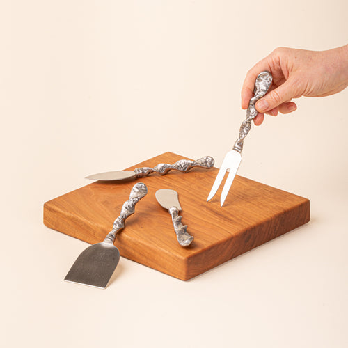Four stainless steel cheese utensils with textured handles arranged on a wooden board, with a hand holding the cheese fork.