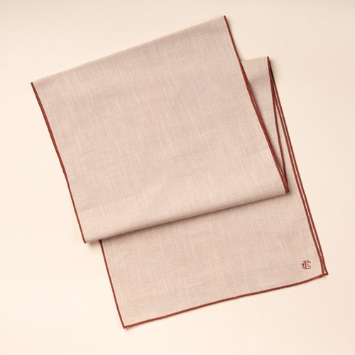 A folded beige table runner with terracotta-colored piping and embroidered monogram.