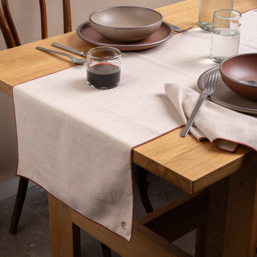 A table set with ceramic pottery, glasses of wine and water, silverware, and a beige table linen with terracotta-colored piping and embroidered monogram.