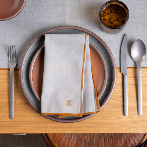 A place setting with a folded light blue table napkin with rust-colored piping and embroidered monogram, with plates in a complementary colorway.