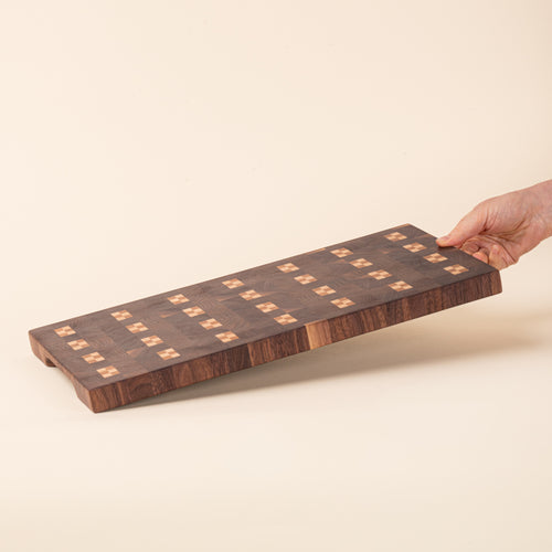 A hand holds out a dark rectangular wooden cutting board with repeating light geometric inlay pattern on a white background.