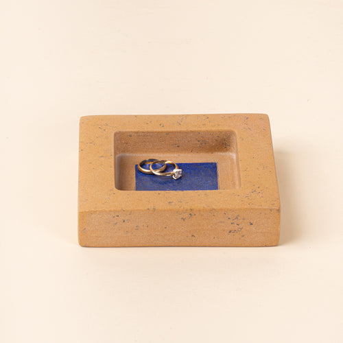 Rectangular tan speckled jewelry dish with a recessed center and blue interior, holding two gold rings.