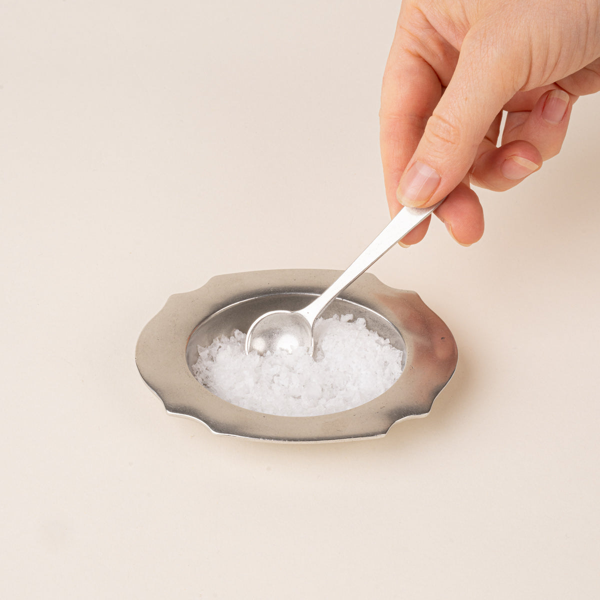 A pewter salt cellar with a broad scalloped rim, filled with salt and a hand holding a small matching serving spoon.