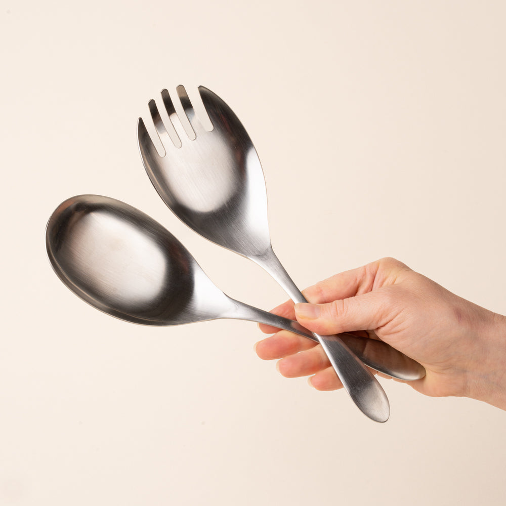 A hand holds a stainless-steel salad serving set with an oversized spoon and four-tined serving fork, featuring smooth oval heads and gently tapered handles, photographed on a warm cream background.