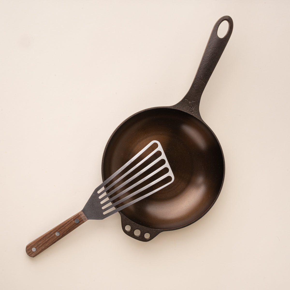 Slotted stainless steel spatula with a wooden handle, resting on a small cast iron pan.