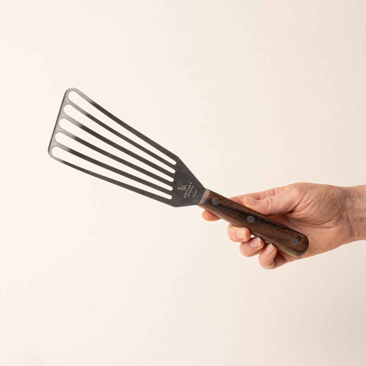 A hand holds a slotted stainless steel spatula with a wooden handle.