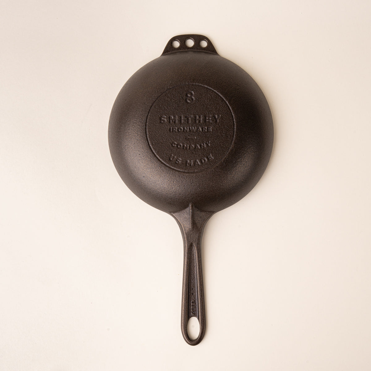 Back of a dark brown small cast iron skillet with a smooth cooking surface and three holes on the opposite side, with a handle and a stamp on the back that reads Smithey Ironware Company.