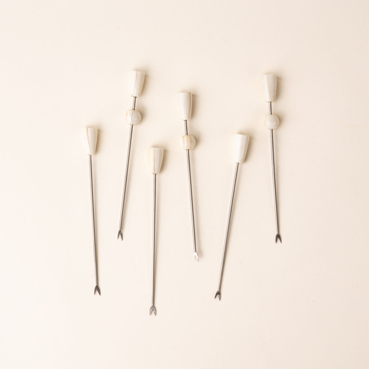 Six slender metal cocktail or appetizer picks with small two-pronged fork tips and sculptural ivory-colored handles, arranged vertically on a warm cream background.