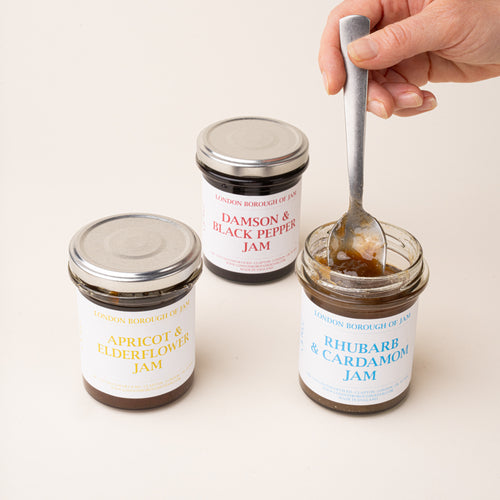 Three glass jars with a white label of Damson & Black Pepper, Apricot & Elderflower, and Rhubarb & Cardamom Jams, with a spoon grabbing some jam