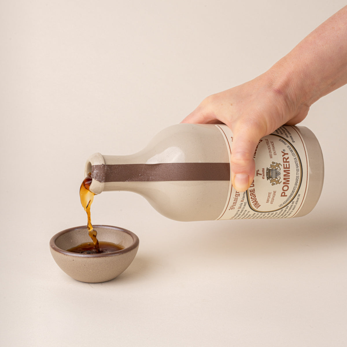 A hand pours Pommery sherry vinegar from a white ceramic bottle with a brown ribbon into a small beige bowl.