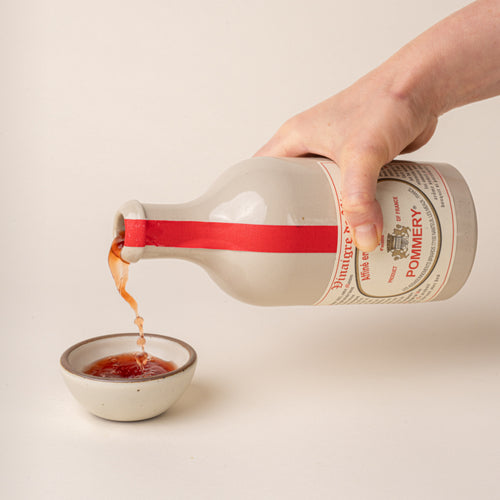 A hand pours Pommery red wine vinegar from a white ceramic bottle with a red ribbon into a small cream-colored bowl.