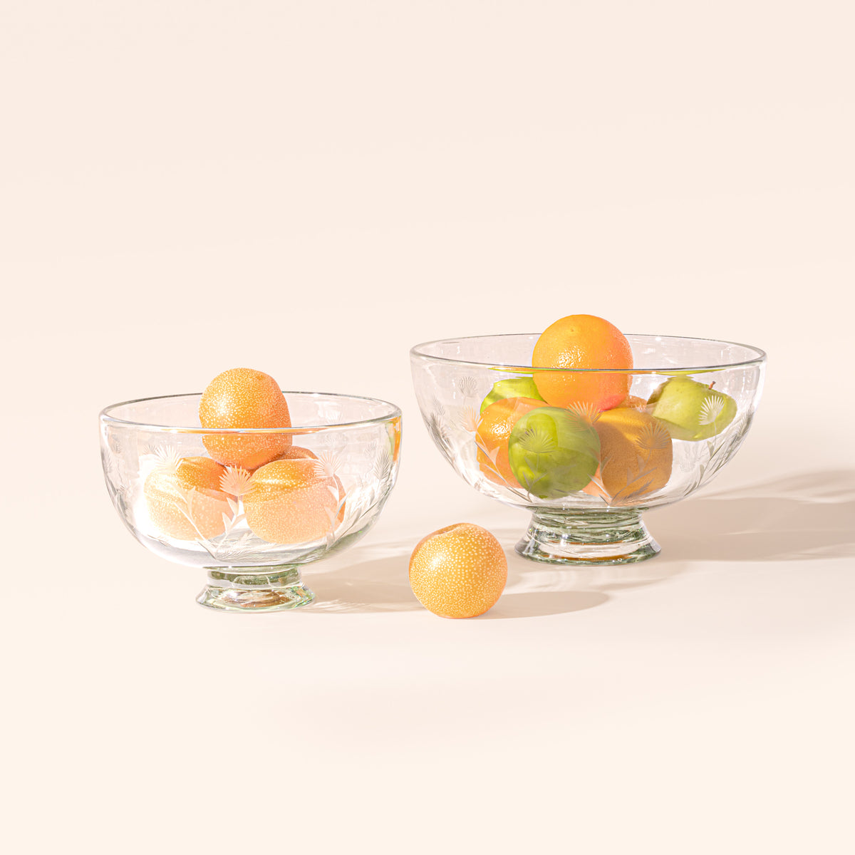 Two clear glass footed bowls in different sizes with etched floral designs, filled with oranges and apples on a cream background.