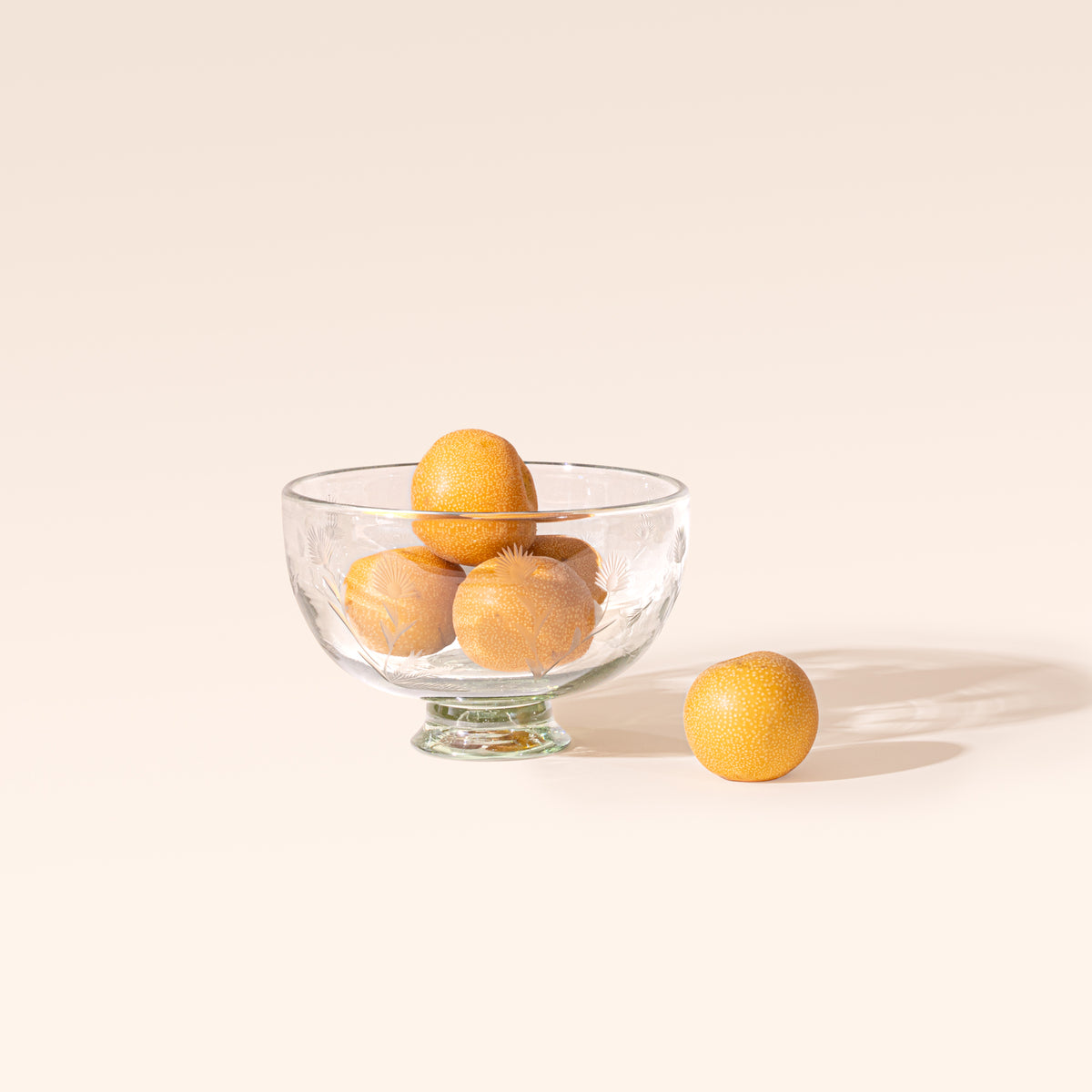 A smaller clear glass footed bowl with etched floral designs, filled with oranges on a cream background.