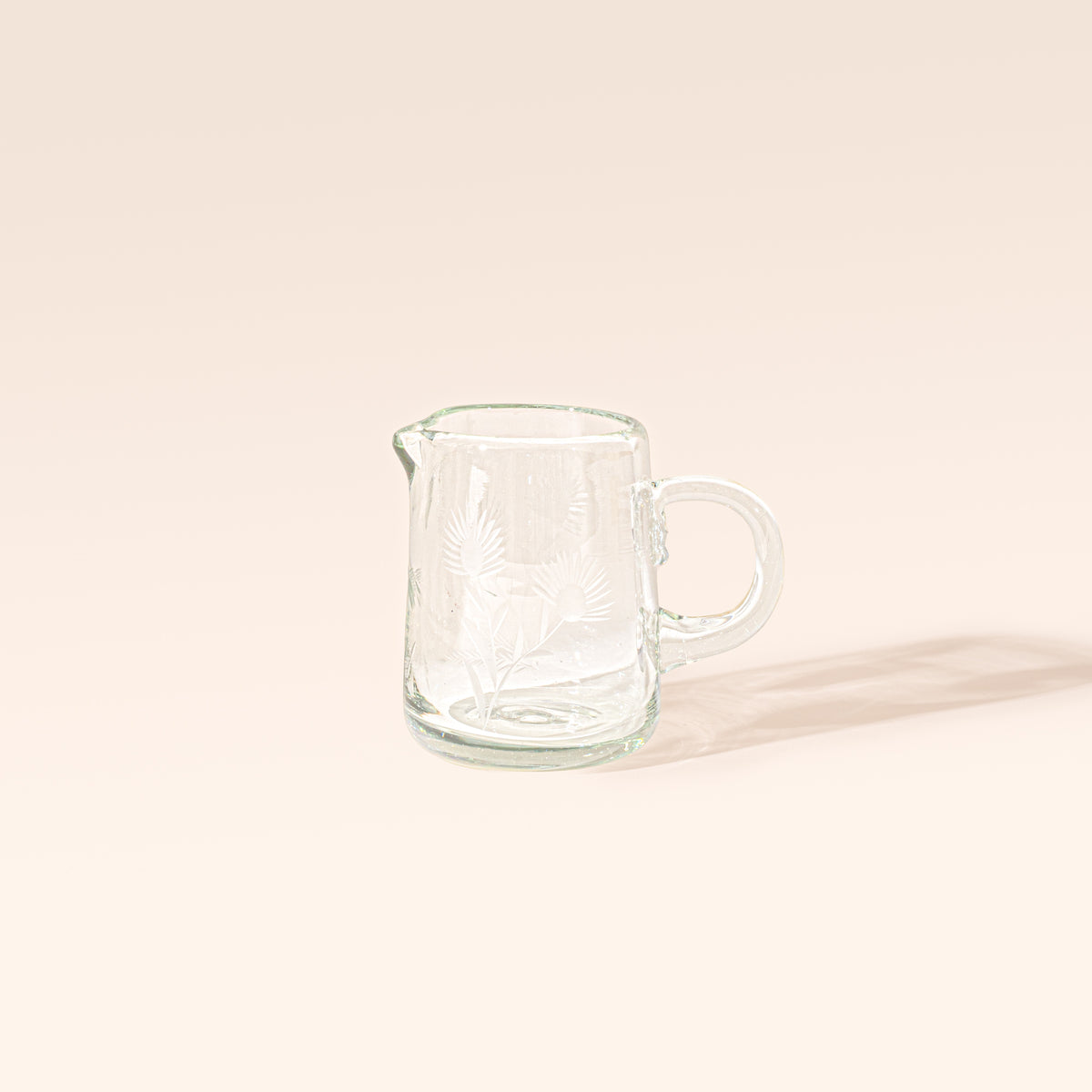 Clear glass creamer with etched floral designs, a rounded handle, and a pouring spout on a cream background.