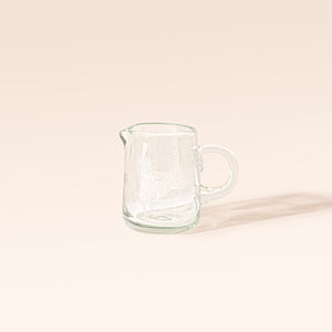 Clear glass creamer with etched floral designs, a rounded handle, and a pouring spout on a cream background.