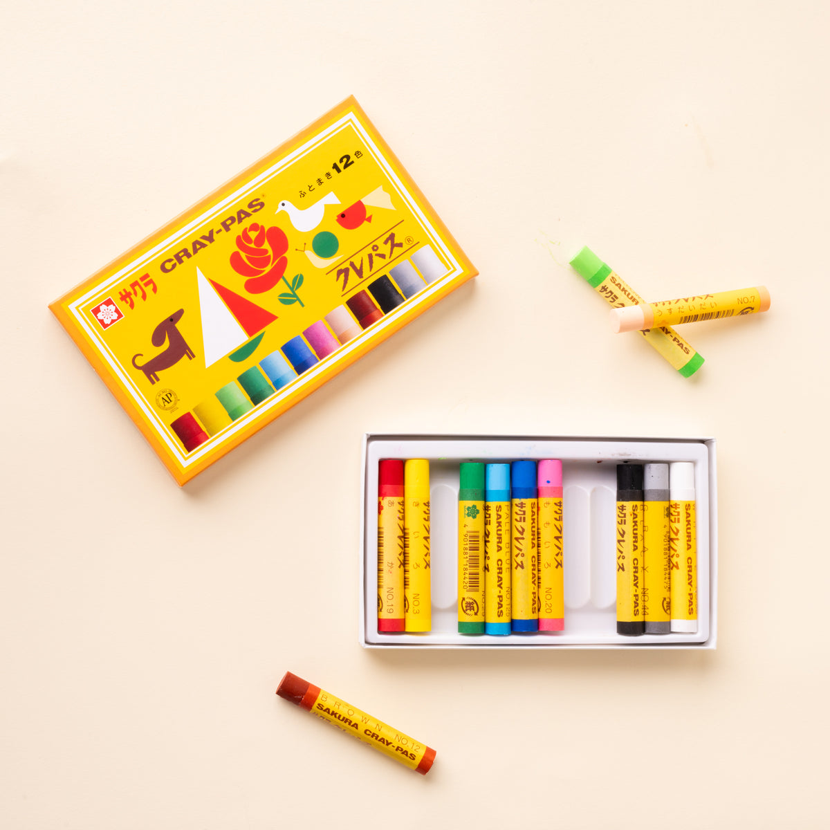 Open box of Sakura Cray-Pas oil pastels with colorful sticks arranged in a tray and four loose pastels beside the illustrated lid.