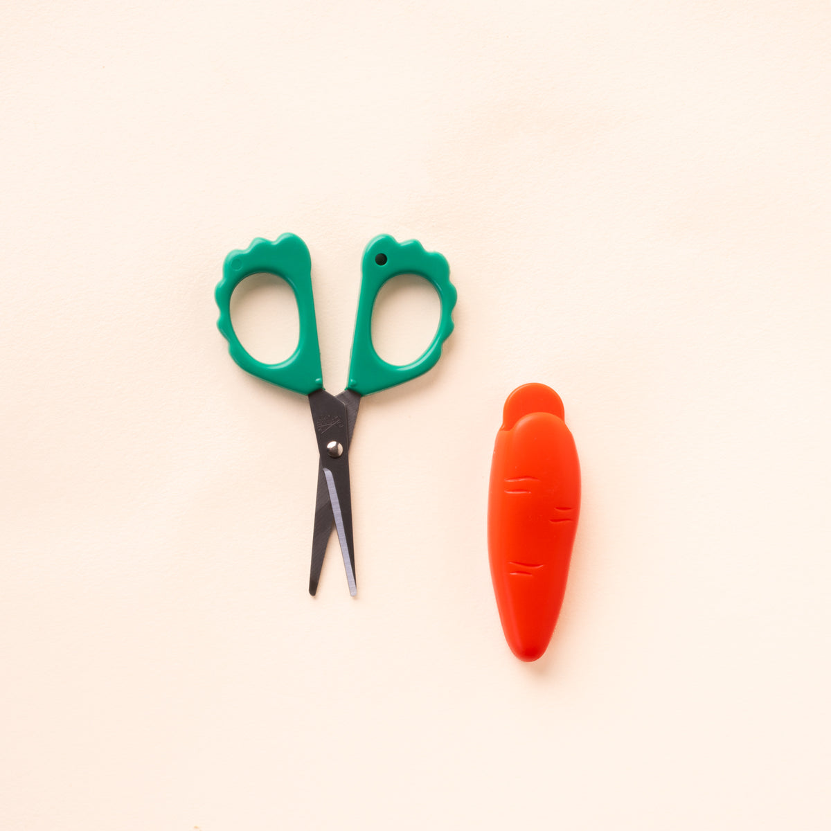 Small scissors with green scalloped handles and black blades, next to an orange carrot-shaped protective cover on a cream background.