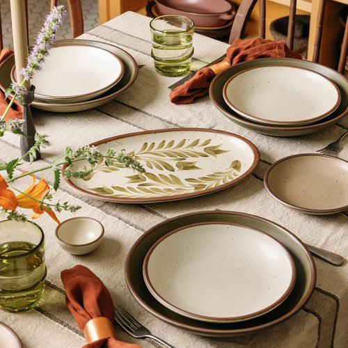 A table set with handcrafted dinnerware, an oval serving platter, green glassware, rust-colored linen napkins, and wildflowers on a striped tablecloth.