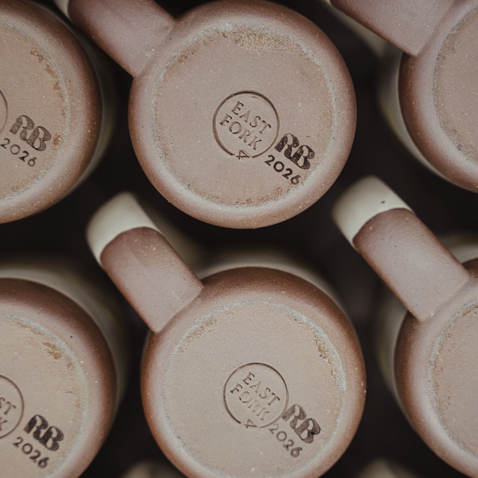 Close-up of multiple ceramic mugs arranged in a grid, showing the bottom of each mug with a stamped logo reading 'EAST FORK RB 2026'. 