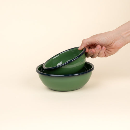 A hand holds a small enamel bowl to nest in a medium enamel bowl; both bowls are green with a dark rim.
