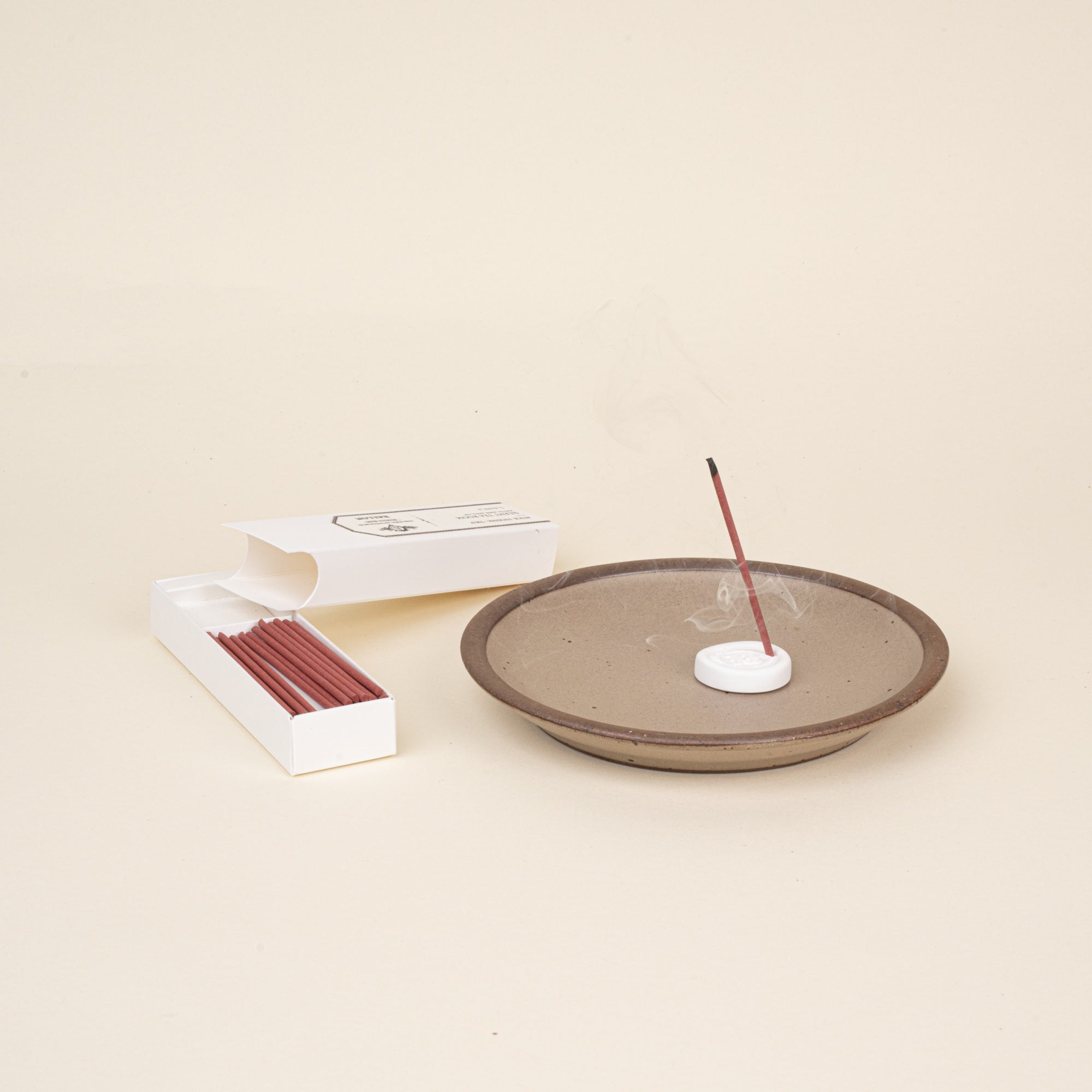 Lit stick incense in a holder on a ceramic dish, with open box of incense sticks beside it.