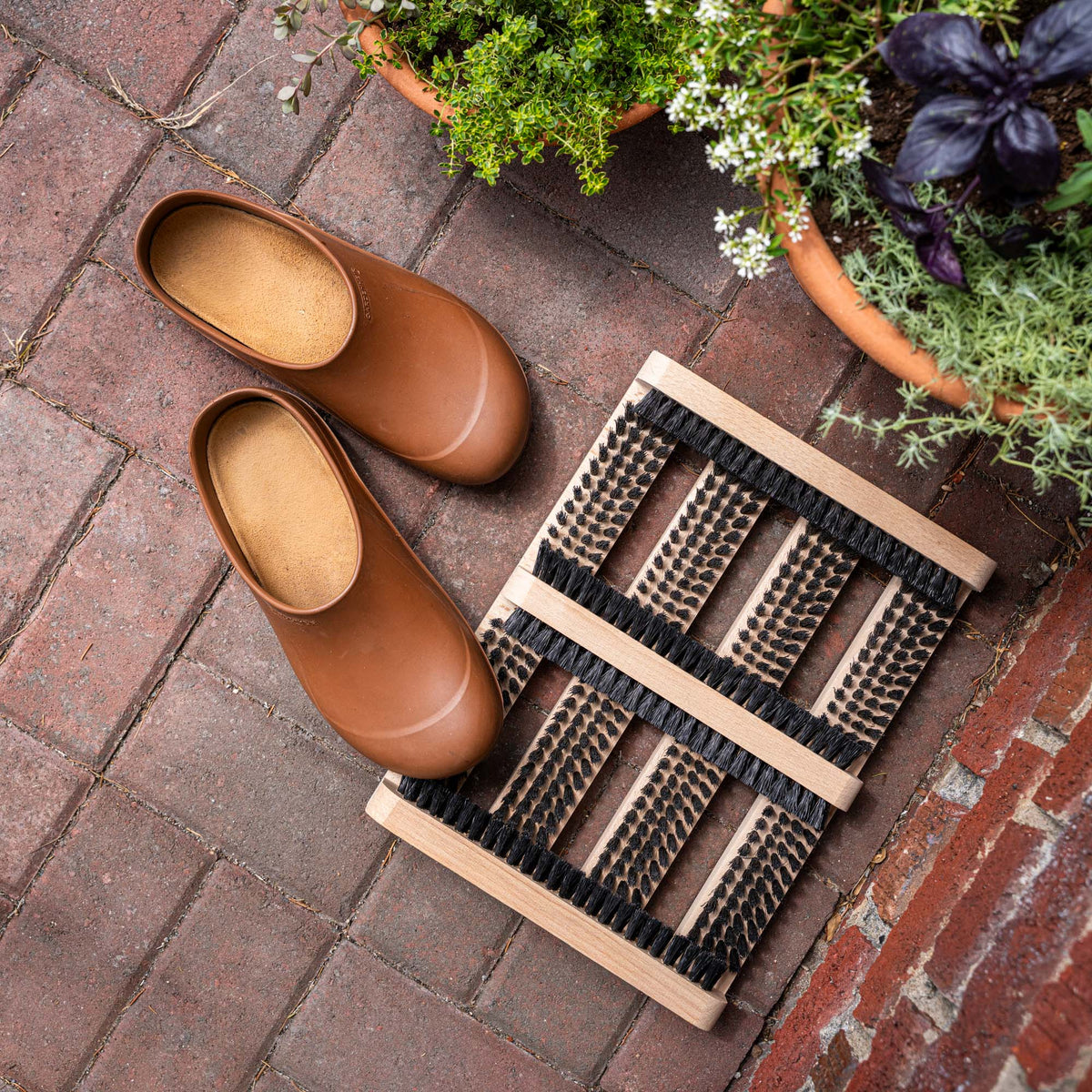 Pair of brown rubber garden clogs beside a wooden boot brush on a brick patio with potted plants.