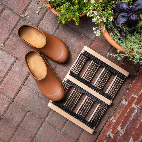 Pair of brown rubber garden clogs beside a wooden boot brush on a brick patio with potted plants.