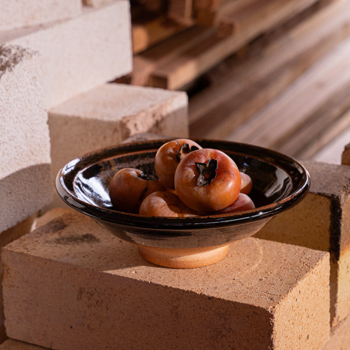 A handmade ceramic bowl with a wood fired earthy muddy glaze and an unglazed base, filled with persimmons in a kiln.