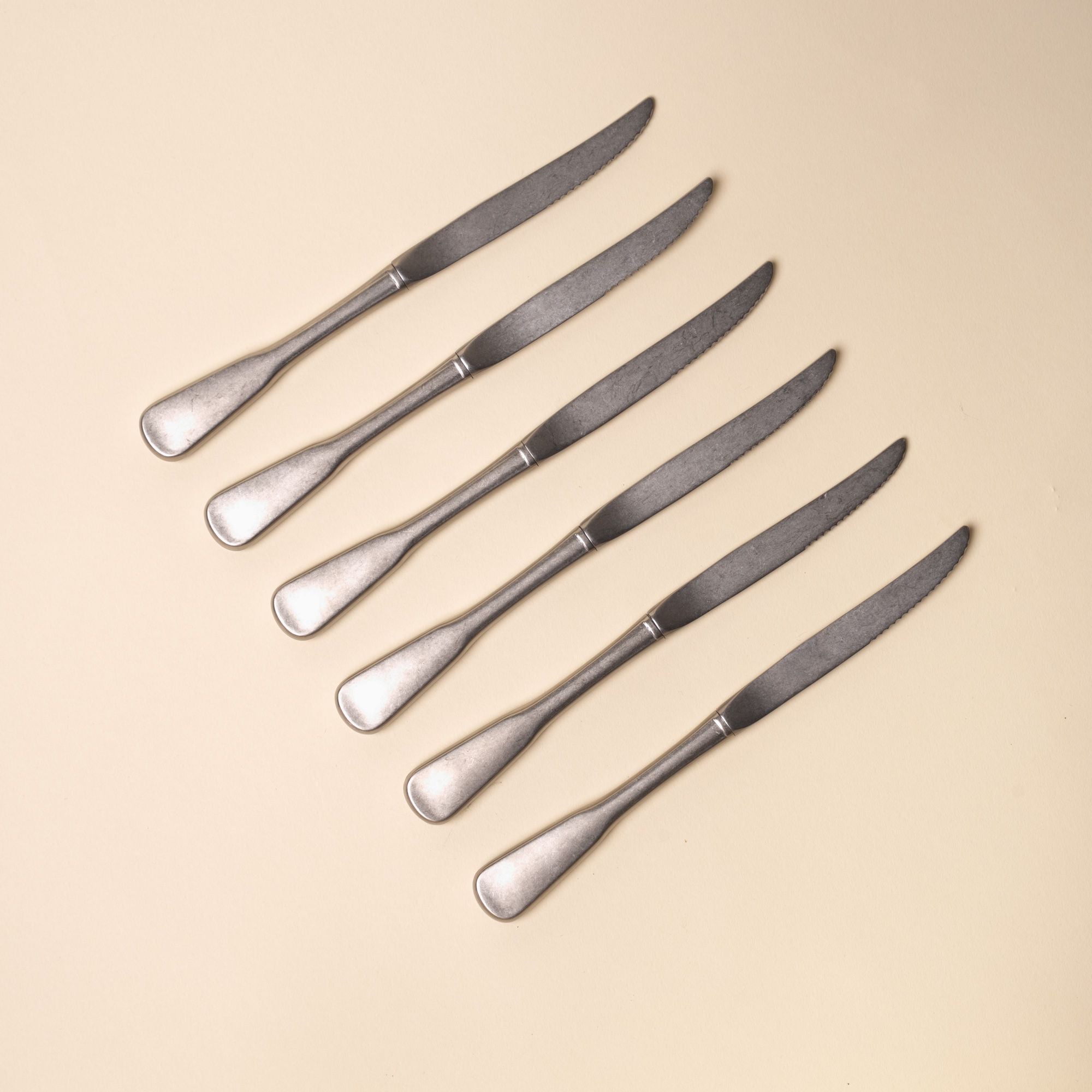 Six matte stainless steel dinner steak knives with rounded handles arranged diagonally on a neutral beige background.