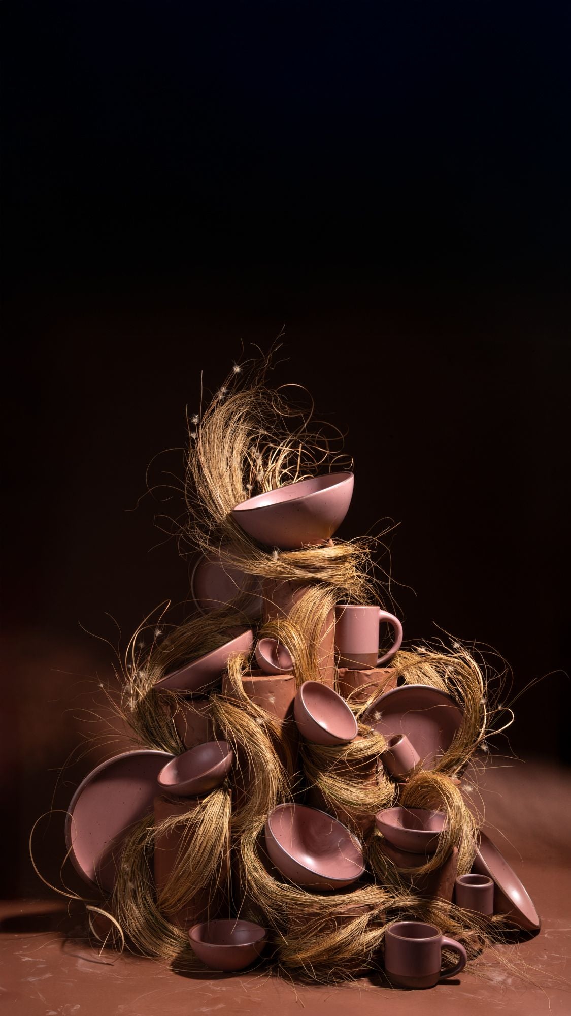 Stack of dusty mauve pink ceramic bowls, plates, and mugs piled together with strands of dried grass against a dark background.