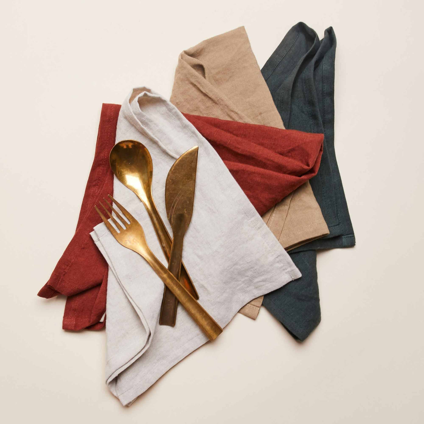 A set of four linen napkins in different colors—beige, red, teal, and white—neatly folded with a gold-colored fork, spoon, and knife placed on top.