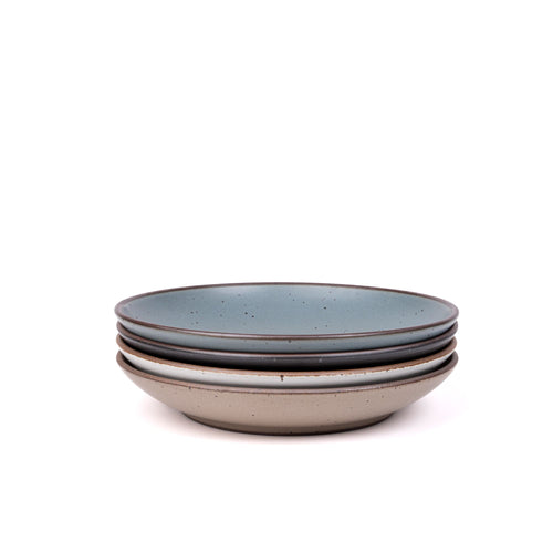 A stack of 4 large ceramic plates with a curved bowl edge in a blue-grey, graphite black, cool white, and warm pale brown colors featuring iron speckles and an unglazed rim.