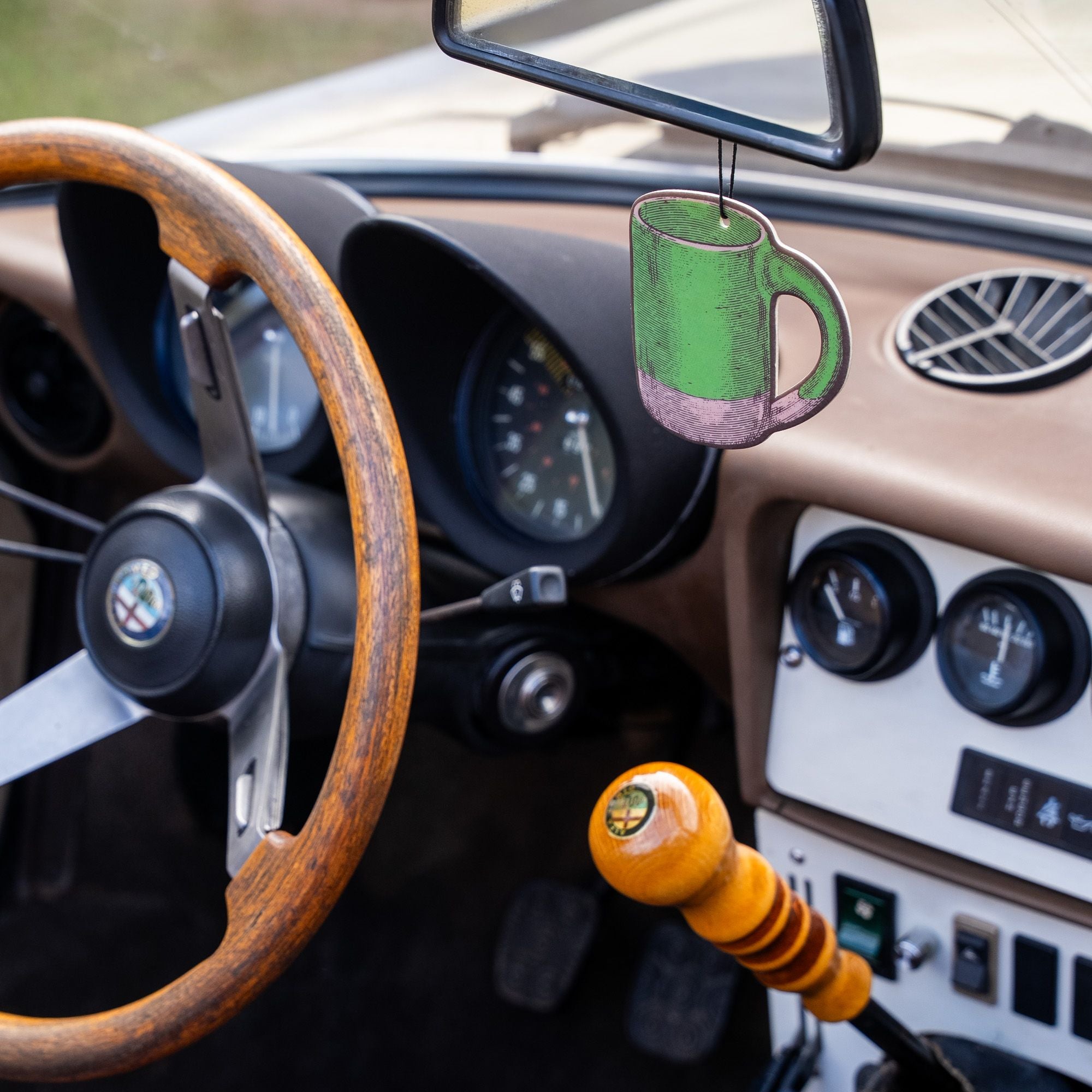 Green mug-shaped East Fork air freshener hanging from a rearview mirror inside a vintage car with a wood steering wheel and classic dashboard.