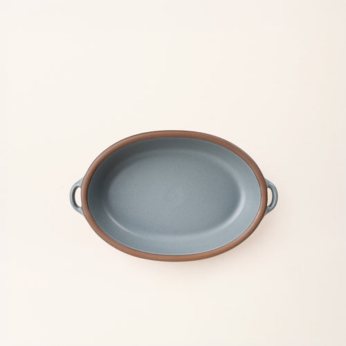 A medium, oval, matte, blue-grey ceramic baking dish with two side handles, on a solid off-white background, photographed from overhead. 
