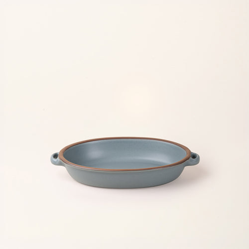 A medium, oval, matte, blue-grey ceramic baking dish with two side handles, on a solid off-white background.