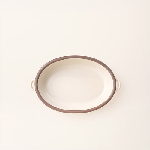 A medium, oval, matte, warm off-white ceramic baking dish with two side handles, on a solid off-white background, photographed from overhead. 
