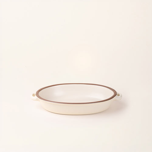 A medium, oval, matte, warm off-white ceramic baking dish with two side handles, on a solid off-white background.