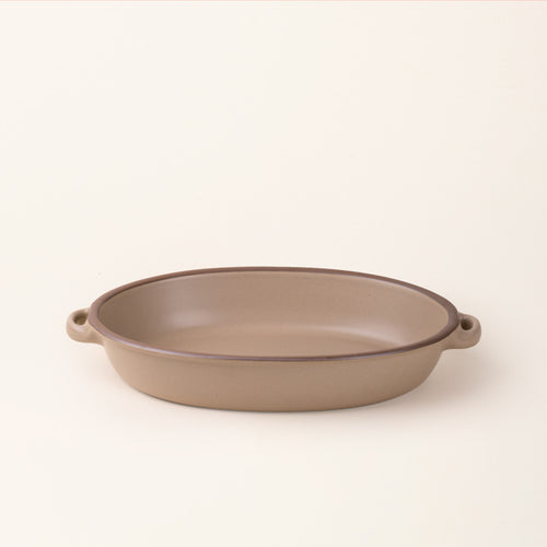 A large, oval, matte, warm pale brown ceramic baking dish with two side handles, on a solid off-white background, photographed from overhead. 